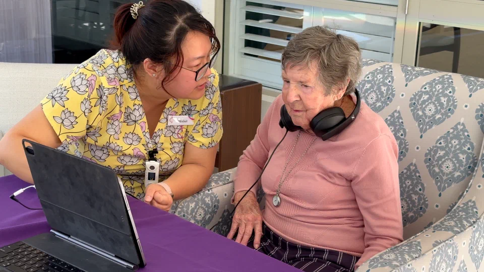 Aged care resident wearing headphones seated alongside a care worker during a Sound Scouts hearing screen.