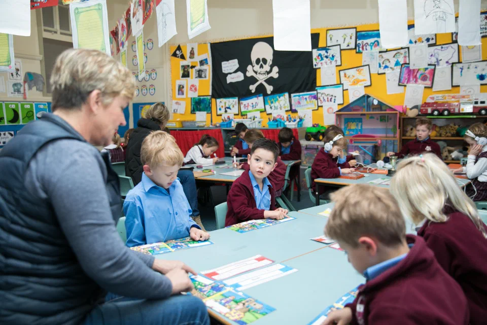 Teacher sitting at the end of a classroom table with 4 students completing a reading activity together.