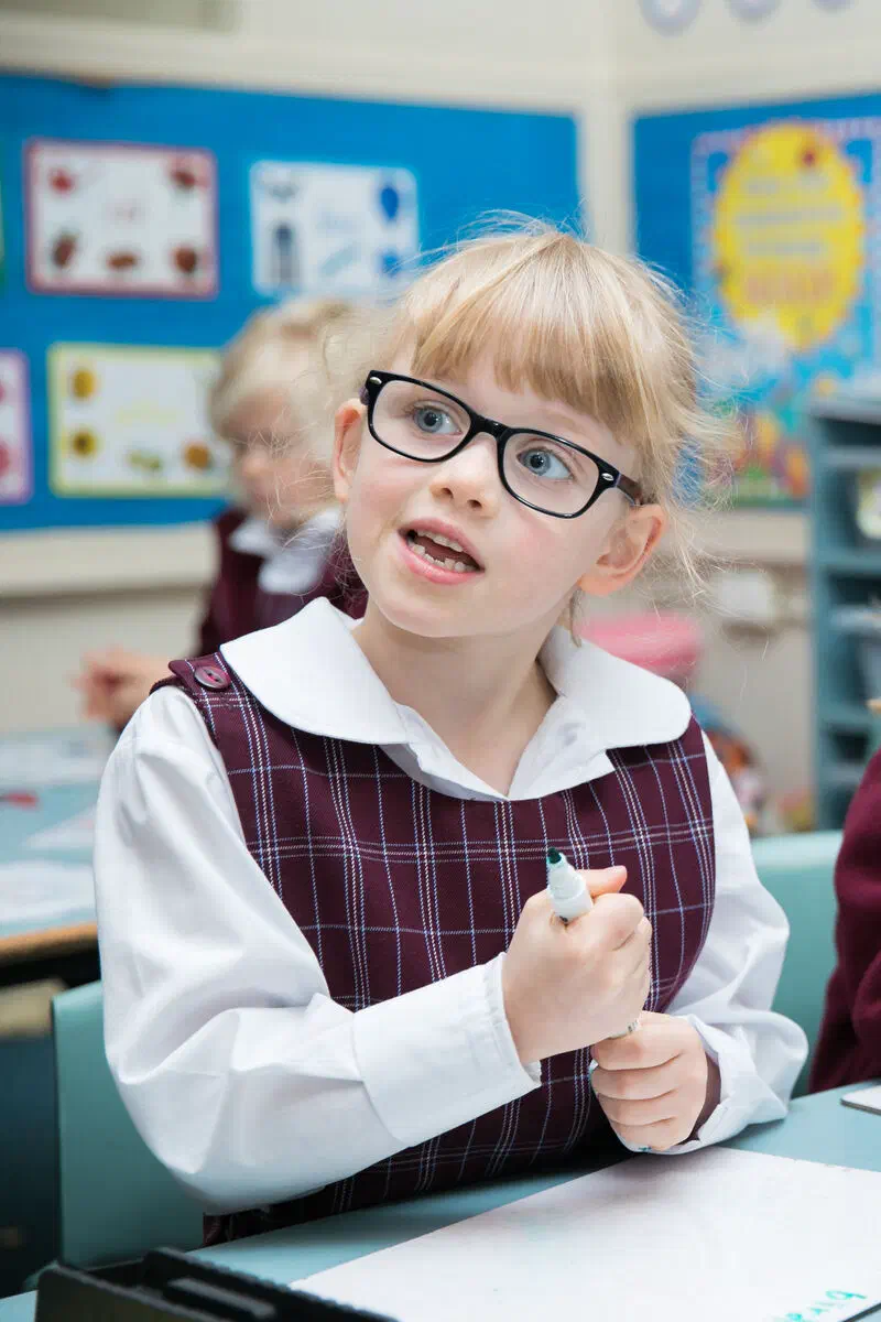 Young student with glasses holding a large pen sitting at a desk.
