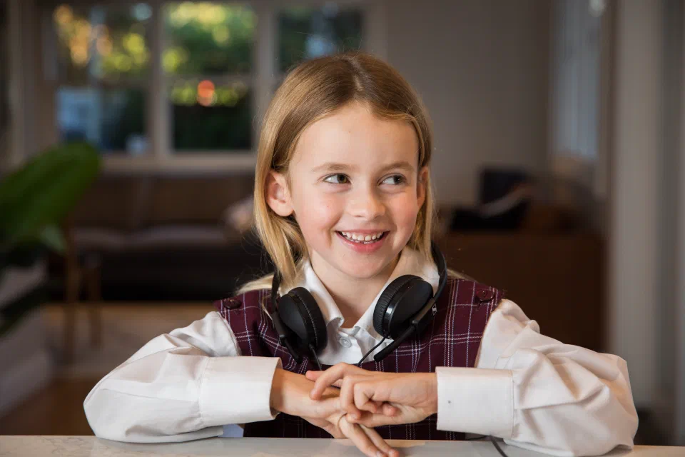 Young girl smiling in school uniform sitting arms folded at a table with headphones around her neck.