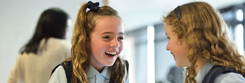Elyse, a young girl in her school uniform, smiling while looking at another young girl in the same uniform.