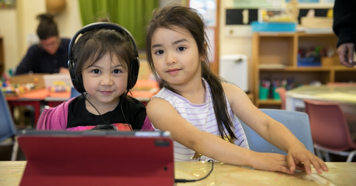 Two girls looking at camera, one with headphones on and tablet in front of her.